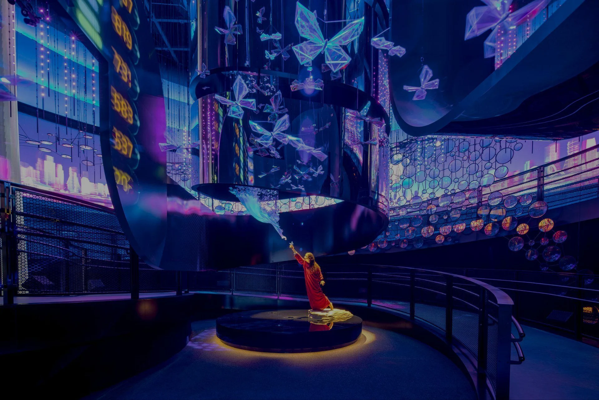 A figure reaches towards suspended butterflies inside Alif, surrounded by sweeping media surfaces and a curving visitor walkway.