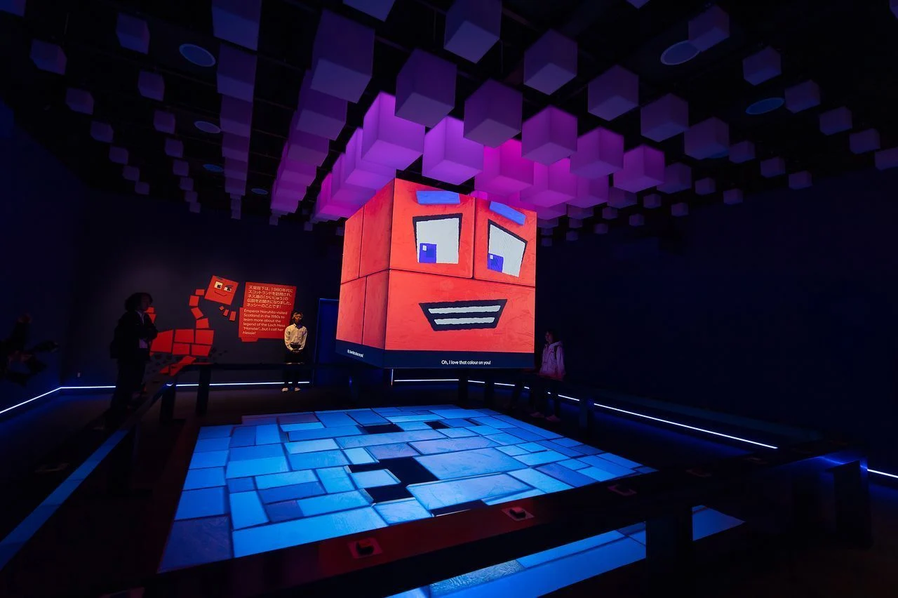 The original PIX character fills an illuminated cube above an interactive floor, with suspended pixel forms extending across the UK Pavilion ceiling.