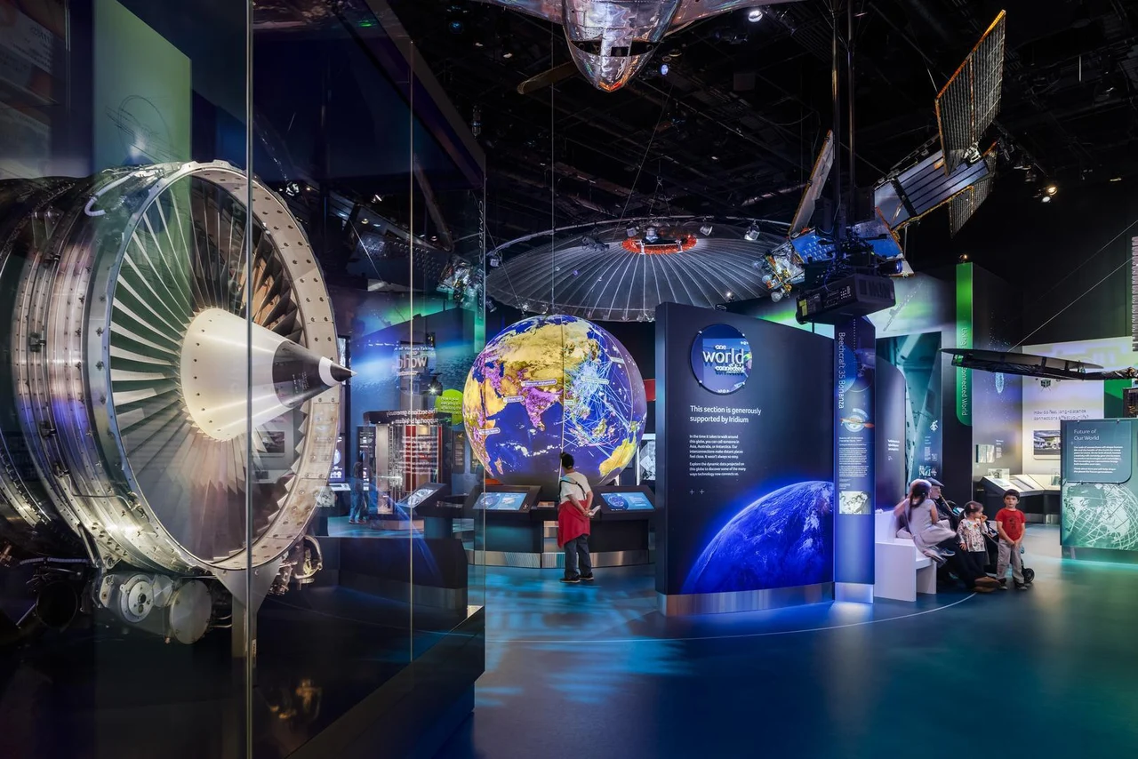 A wide view of the Smithsonian’s One World Connected gallery, with a luminous globe, suspended satellite models and visitors exploring digital displays.